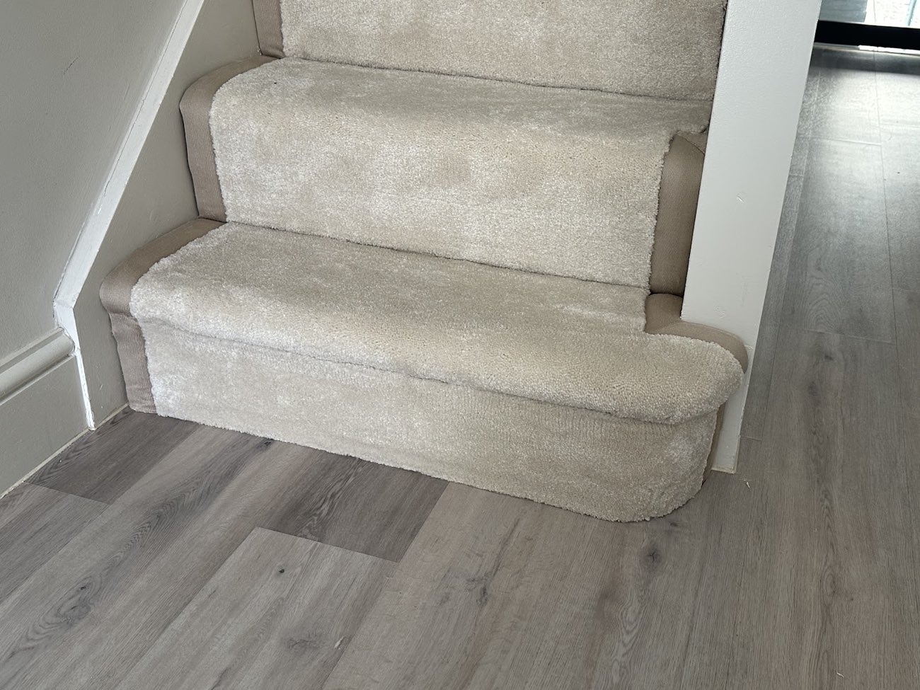 A beige carpeted staircase meeting grey wood-effect vinyl plank flooring in a hallway, with a clean join at the bottom step.